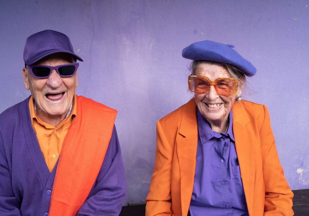 Two funky elderly people wearing bright purple and orange on a purple background