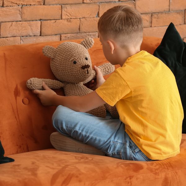 A young boy with his back to the camera sitting on a couch playing with a teddy bear