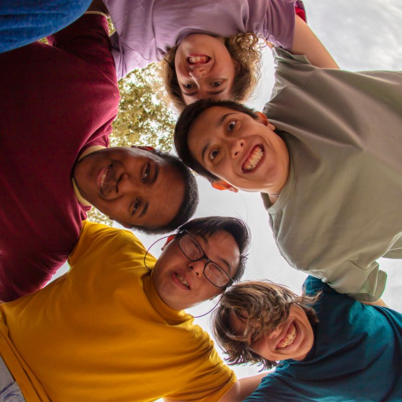 A group of teenagers with intellectual disabilities in a circle embrace looking down at the camera