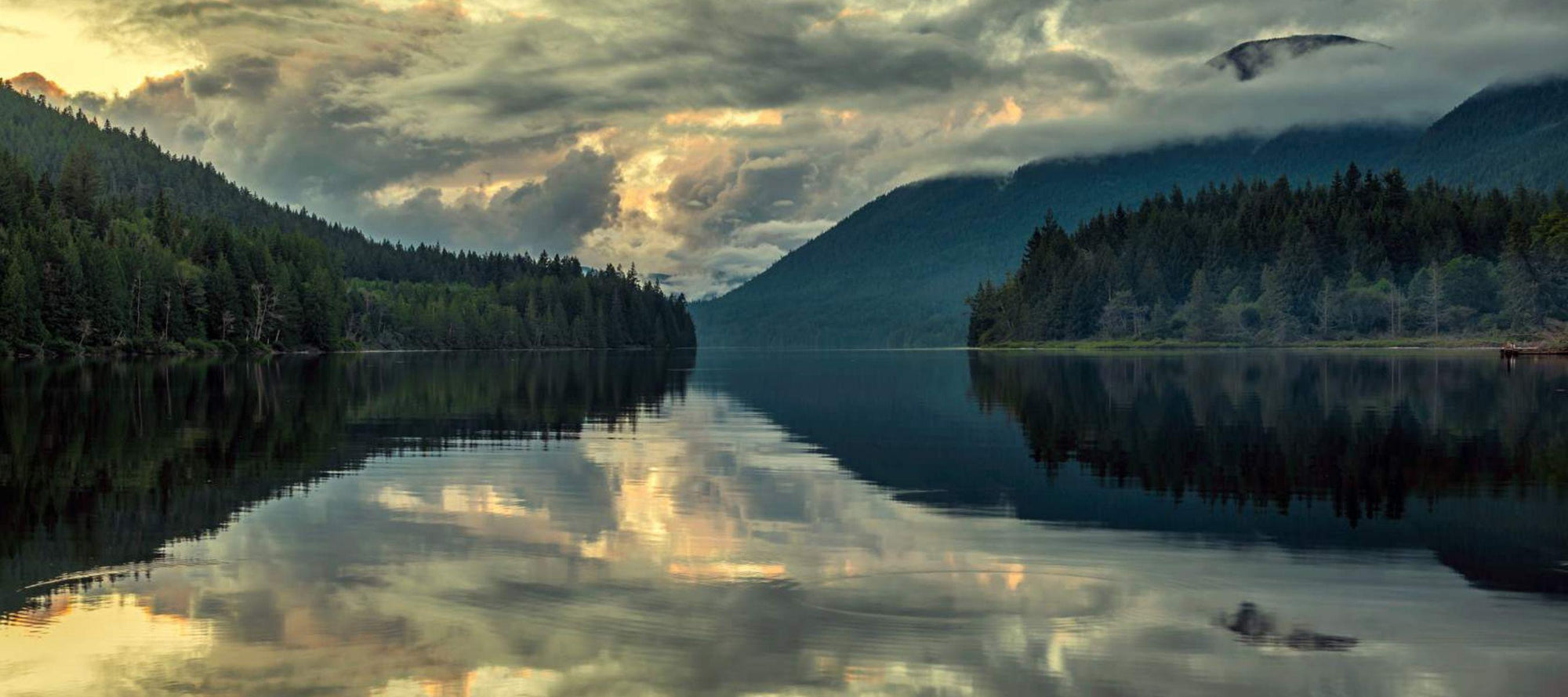 Reflective lake surrounded by forested hills under a cloudy sky