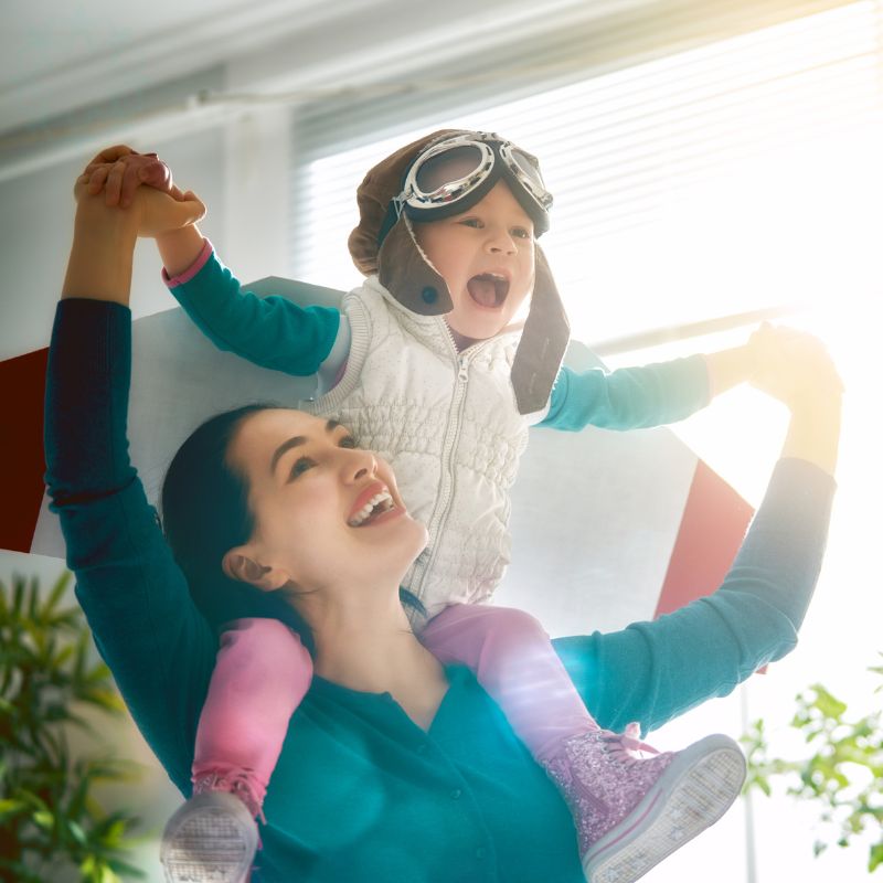 Image of a woman holding a child on her shoulders. The child is wearing aviator goggles and fake wings. 