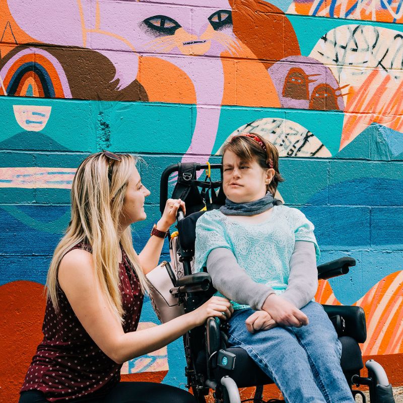 An inclusion employee in front of a colourful wall with an individual in a wheelchair
