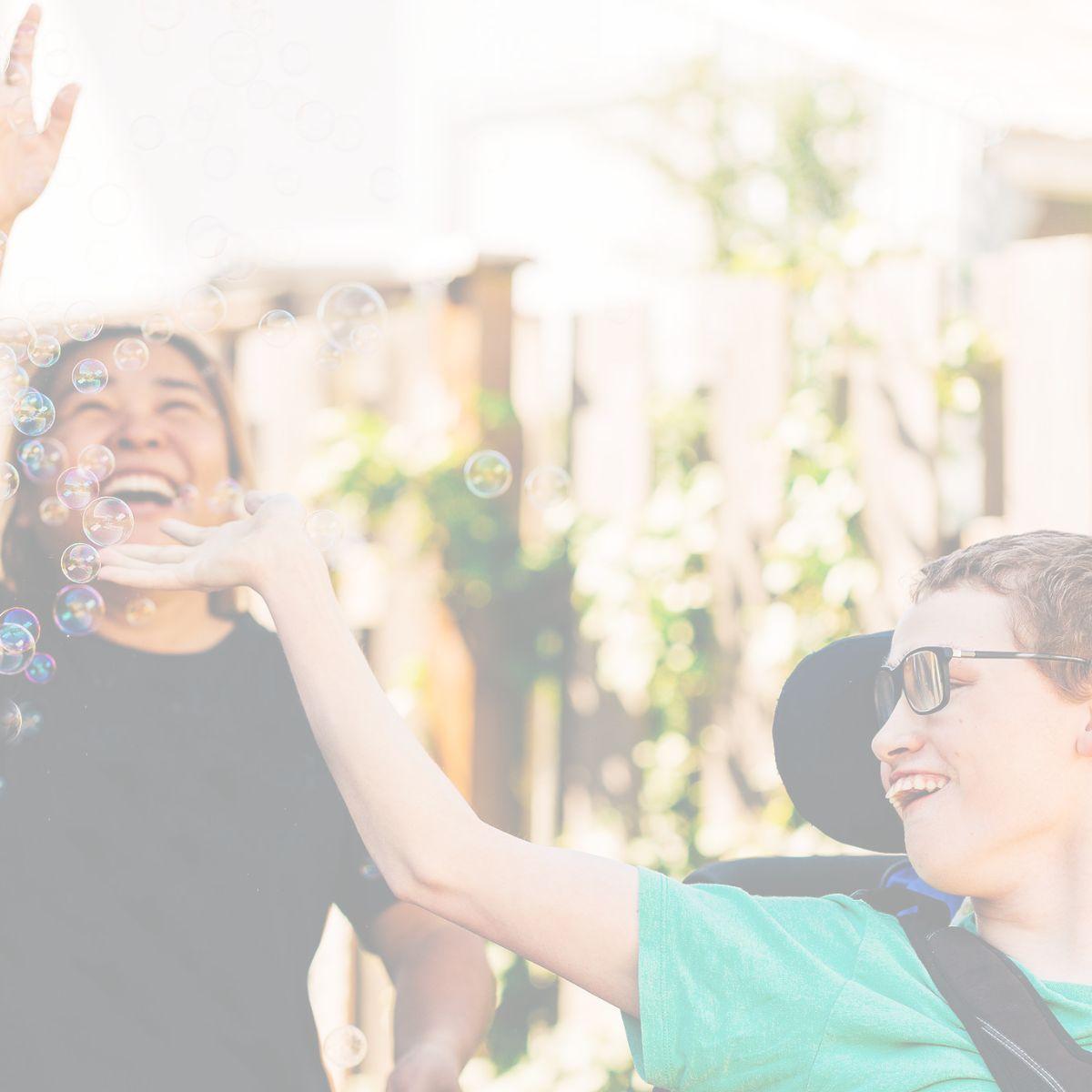 A woman and a young man in a wheelchair catching bubbles in the air. 