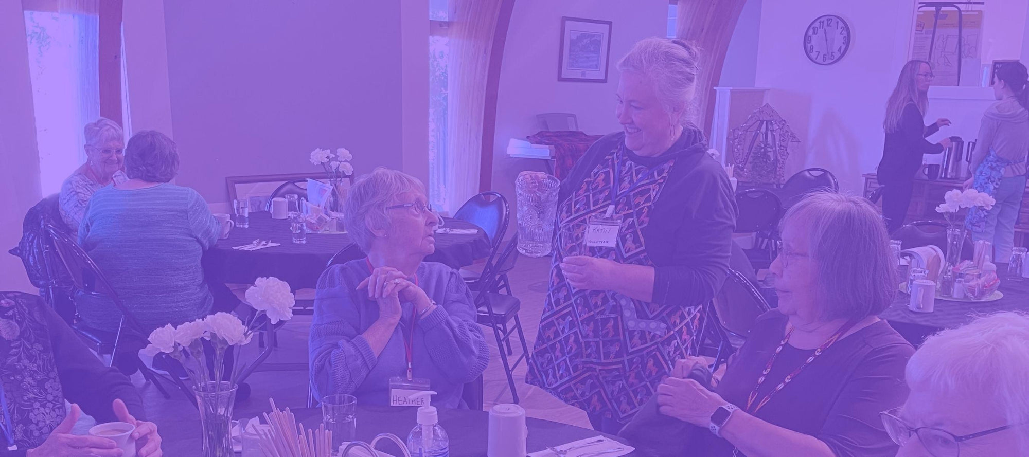 A woman wearing an apron pouring water into a glass for an older woman sitting at a table with her friends
