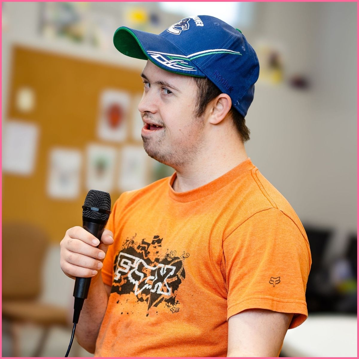 A man in an orange t-shirt and blue hat is singing karaoke. 