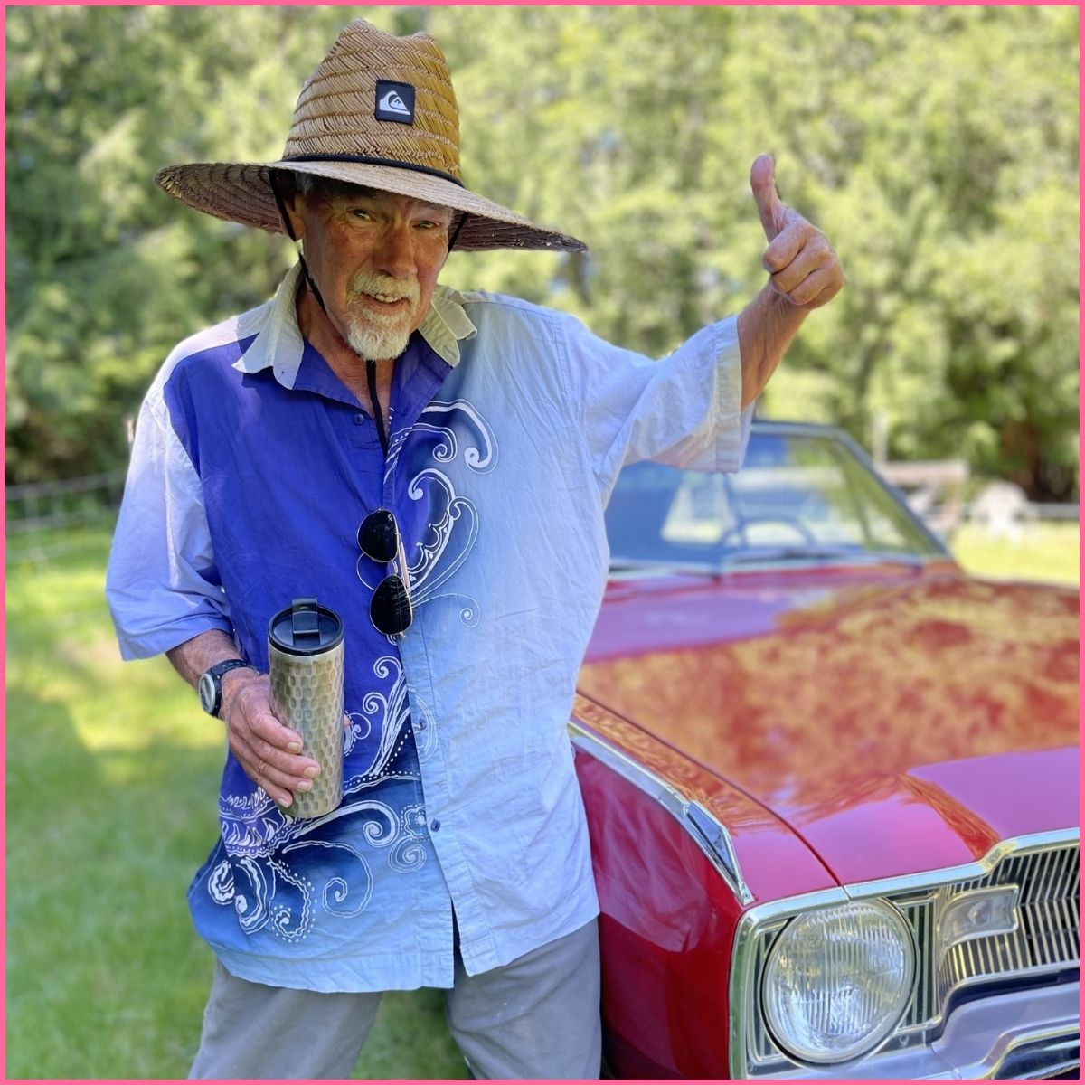 A man with a goatee and sunhat is posing in front of an old, red sports car. 