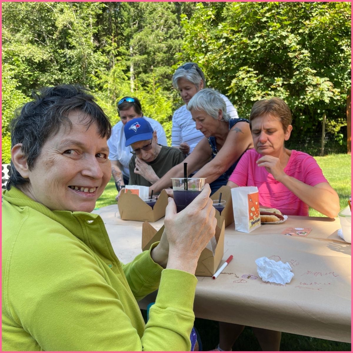 Image of some people sitting at a picnic table doing art on brown craft paper. One woman is turned towards the camera, holding up her drink, cheering the camera. 