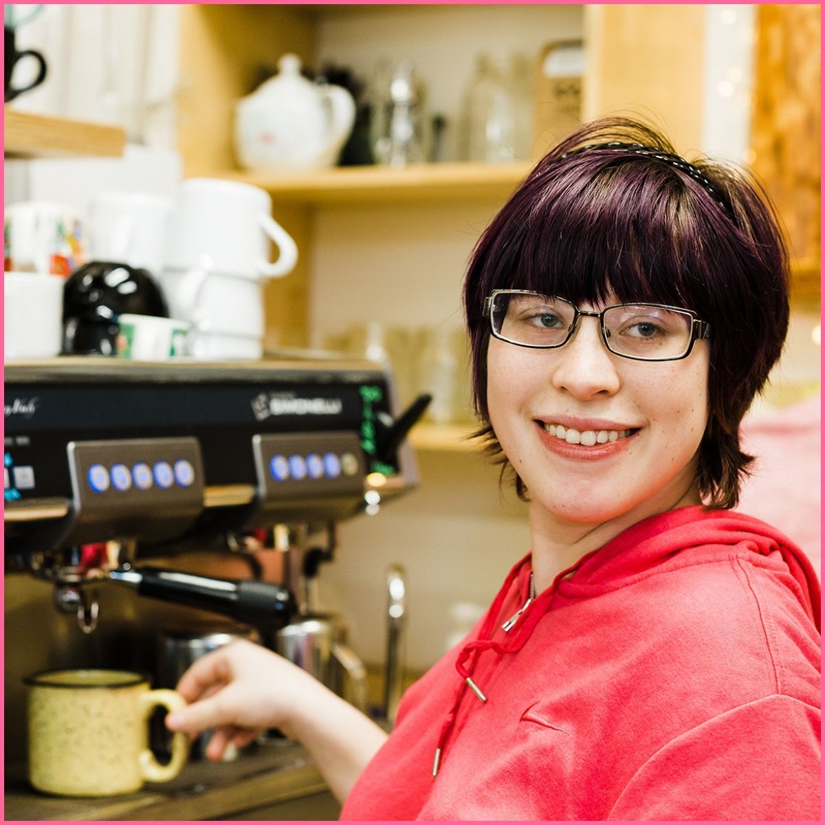 A woman in a red shirt and dark glasses is making coffee at a coffee shop.